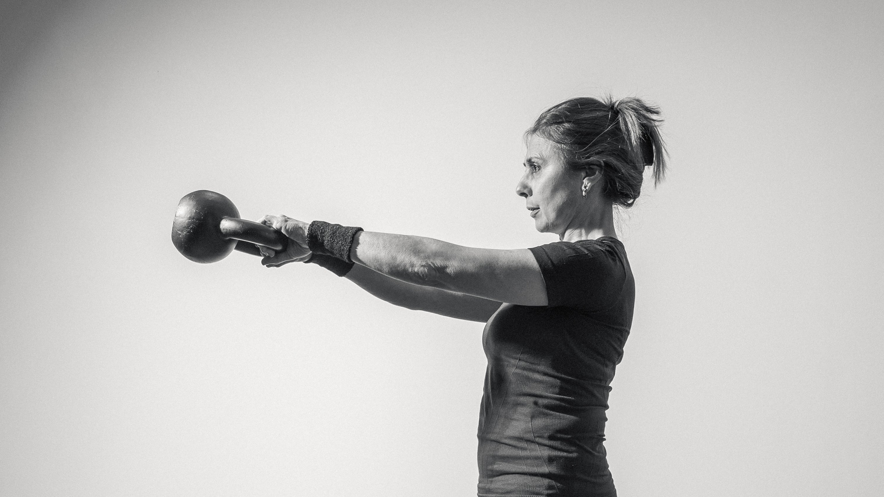 Frau mittleren Alters trainiert mit ausgestreckten Armen und einer Kettlebell, in schwarz-weiß aufgenommen.